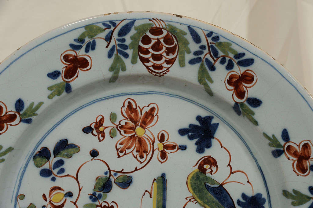 We are pleased to offer this antique Delft charger with parrot is a delightful example of 18th century Lambeth English Delft. The parrot is resting on a branch at one side of a lush garden with oversized flowers. The scene is surrounded by a border