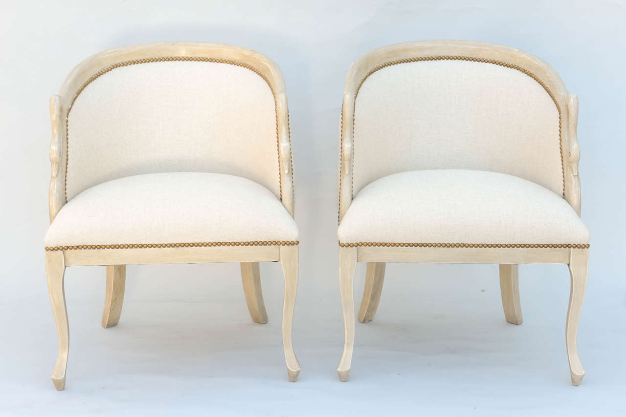 Pair of bergeres, having a painted finish, each barrel form chair with curved crestrail sloping into arms, continuing into carved swan neck terminals; padded back and crown seat upholstered in linen with nailheads, raised on square section stylized