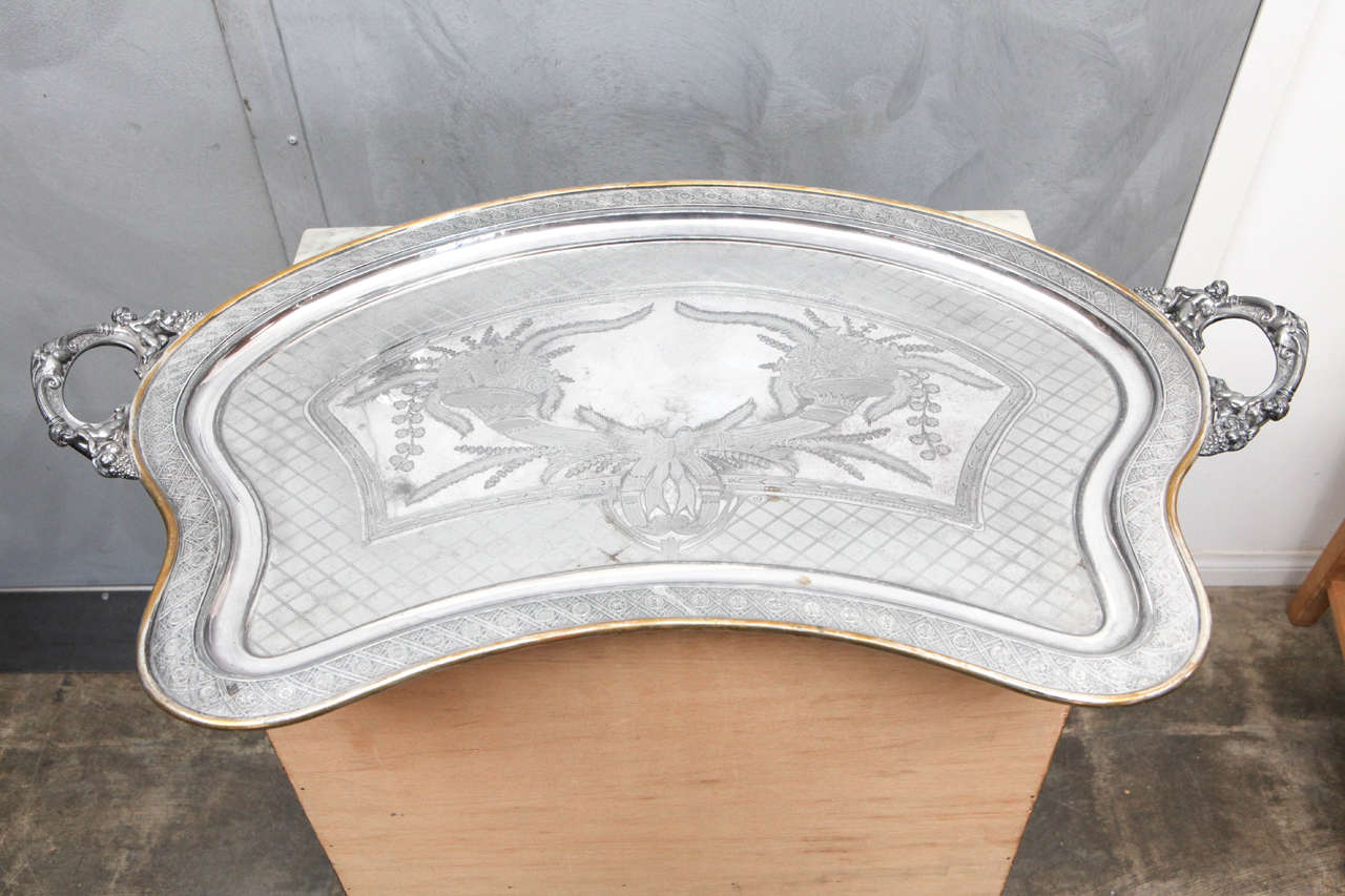 This tray is what is termed a butler tray because it is shaped to fit against the body with two handles. The silver plated tray is beautifully engraved with cornucopias dripping with flora, fauna and fruit. Ribbon bows and a trellis pattern add to