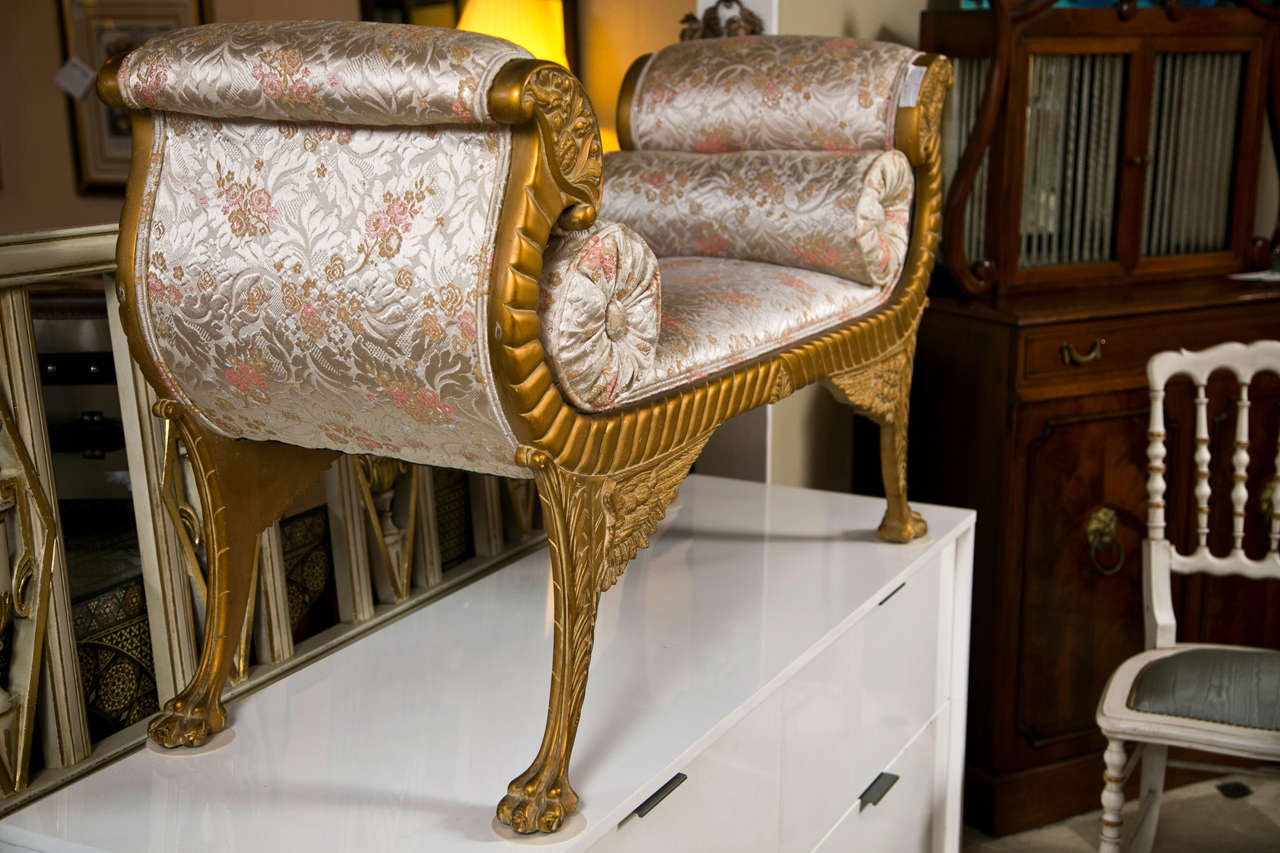 A giltwood bench in the style of French neoclassical. Slightly distressed finish, rolled arms depicting ram's head motif, upholstered in satin, comes with two bolster pillows, raised on winged splayed legs ending in claw feet.