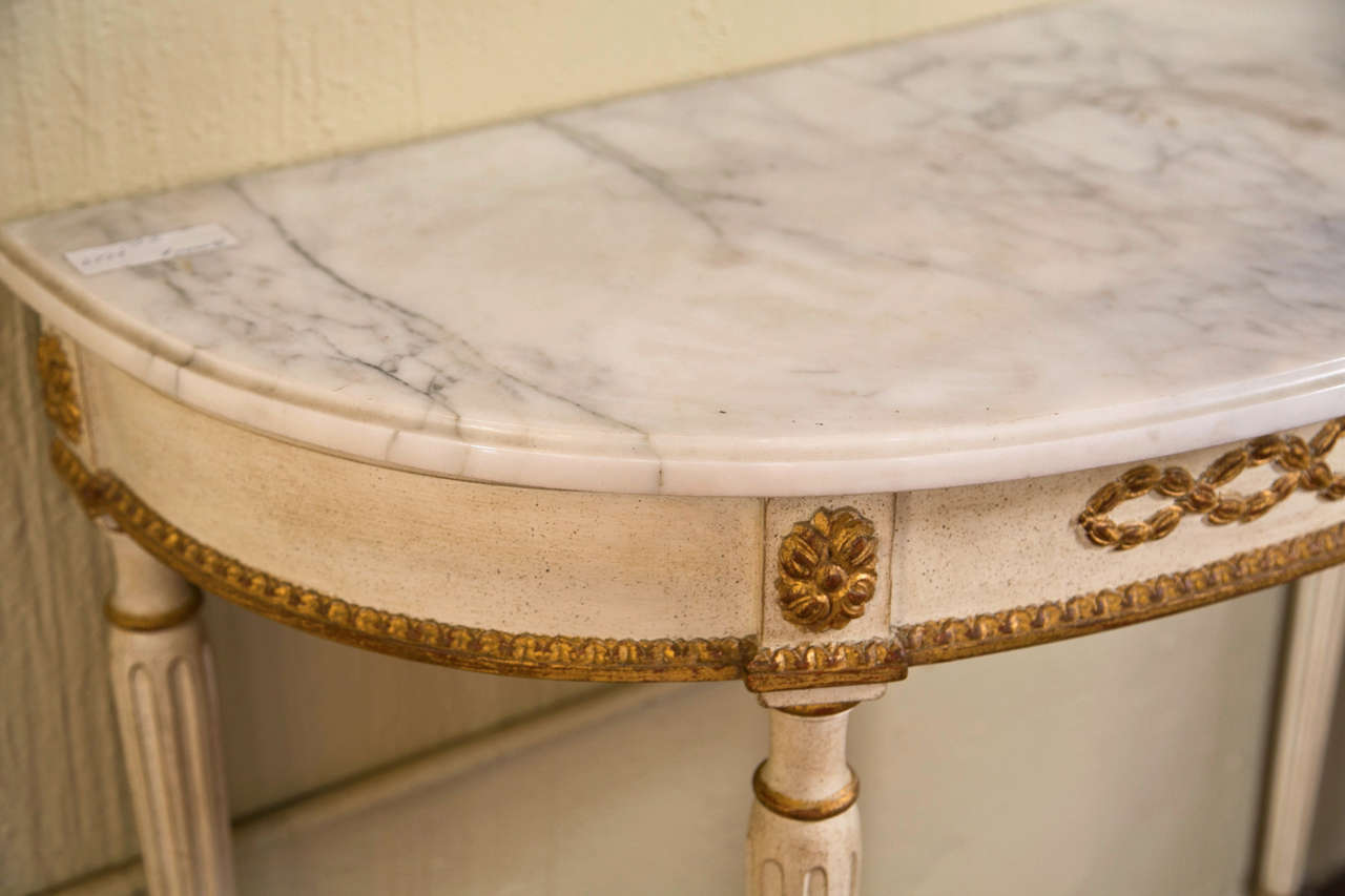 The half-moon white marble atop a painted and parcel-gilt narrow frieze decorated with patera and ribbons, raised on four fluted tapering legs ending in toupie feet.