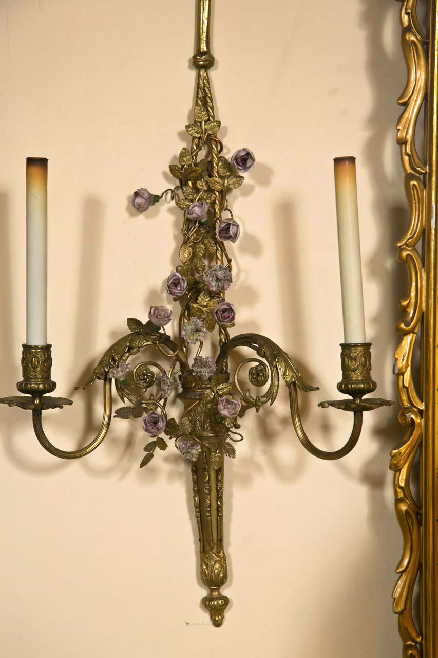 French Belle Époque style brass wall sconces, circa 1930s, the top headed by a ribbon extending to a shaft decorated with porcelain Meissien. Florette and foliate, two-arms each has a long candlestick sleeve.