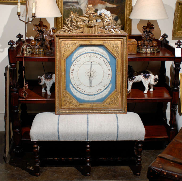 This French Empire style barometer with blue face background, circa 1870 features a rectangular giltwood frame topped with an exquisitely carved crest. A central eagle is flanked with trophies of arms and floral ornaments. The barometer face is