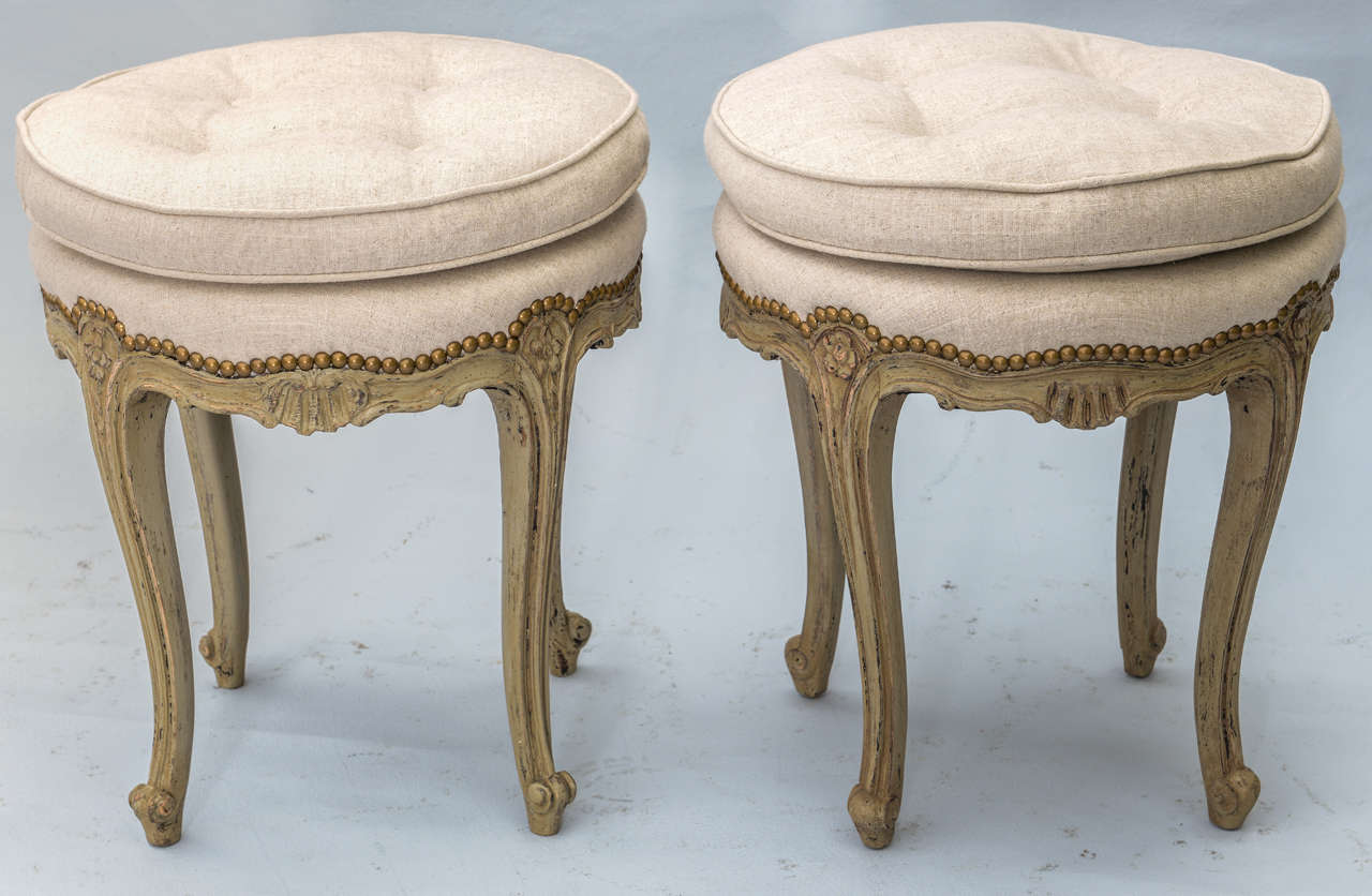 Pair of stools, each tufted round top semi attached cushion on matching deck with nailheads, painted frame with natural wear,  channelled serpentine apron carved with combing, raised on rosette-headed cabriole legs, terminating in scroll