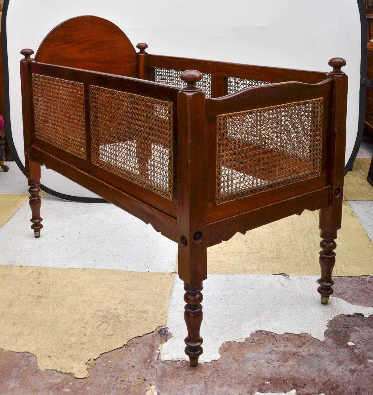 English early Victorian cane sided crib. The head board end is mahogany with a arch top. Each corner is capped with a knob shaped finial. The legs are of a turned a balustrade form with brass caps. Bottom end rails have a scalloped design. The caned