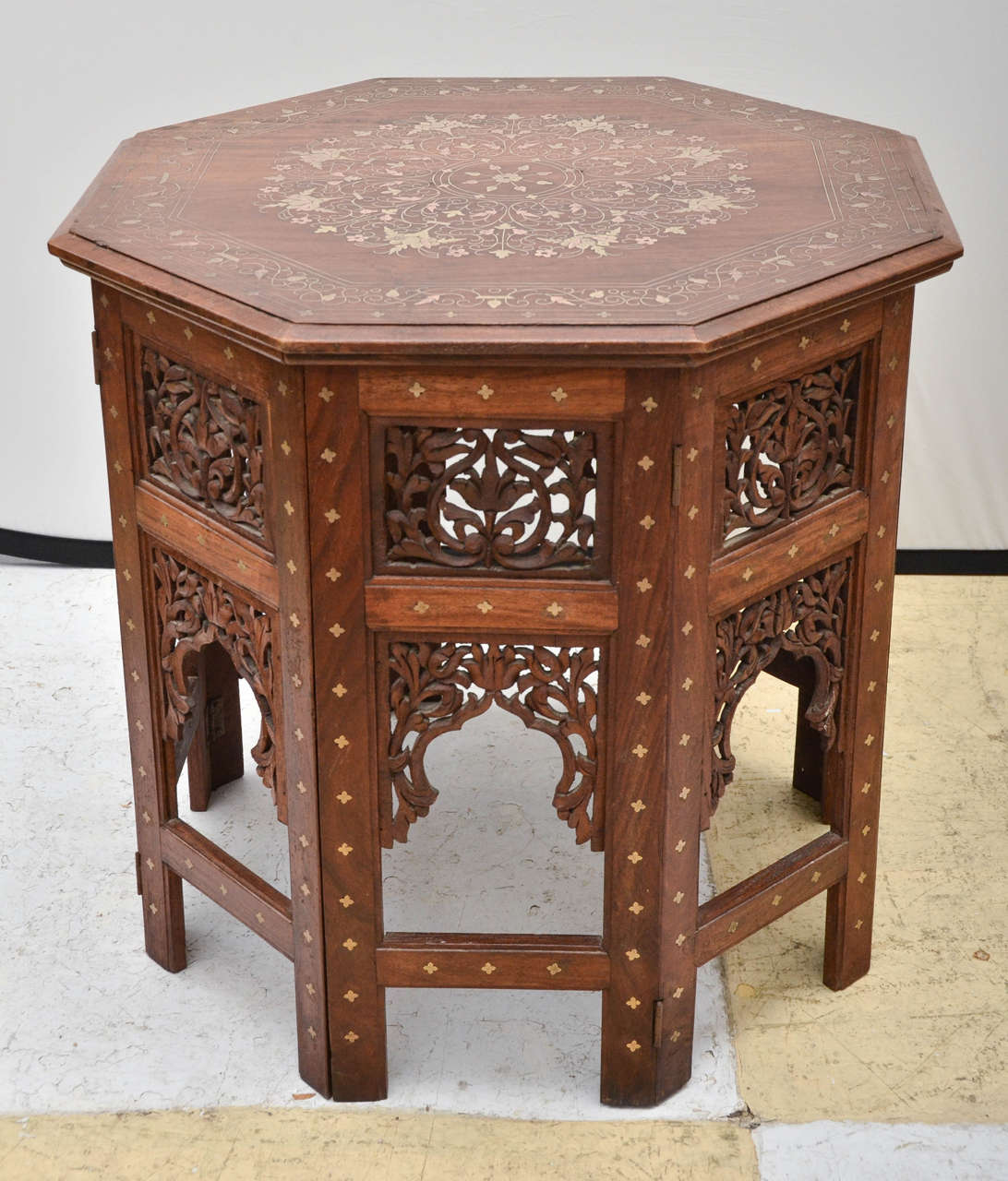 Large octagonal Indian teak occasional table with brass inlay. On a folding pierced fretwork design base. The top is inlaid with brass floral bouquets and trailing vines. A few of the inlays are copper. The sides have cut-out floral designs and