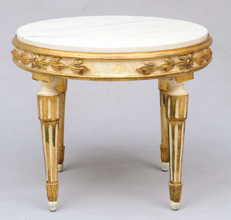 Round accent table, having white veined marble top inset in painted and parcel giltwood frame, its apron carved with foliate and laurel leaf details; raised on round fluted legs