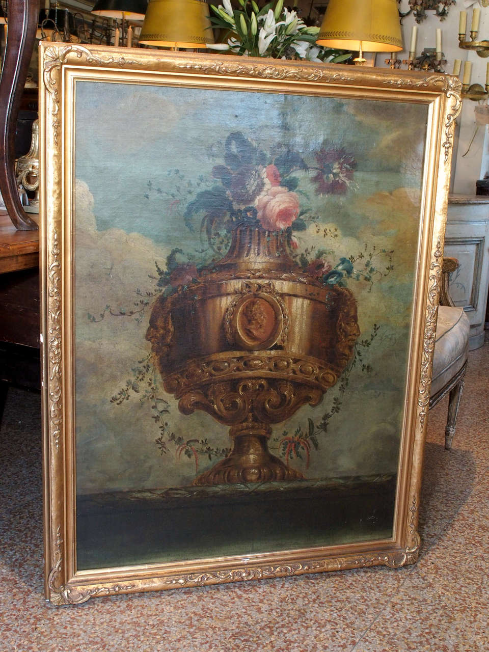 A large framed oil on canvas depicting a carved urn with horned masks with a central medallion in an oval cartouche. Flowers and foliage emerge from the top of the urn.