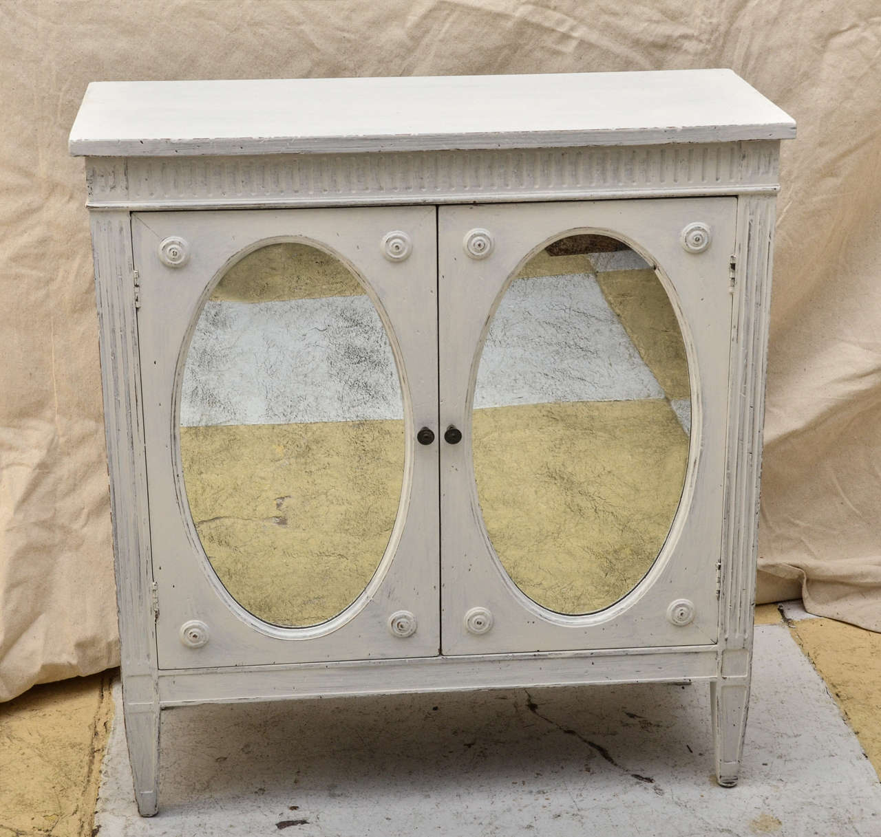 Gustavian Style Pale Grey/White Painted Antiqued Oval Paneled Doors With Inset Antiqued Mirrors. Each Doors Has Round Turned Discs In The Corners. The Top Frieze & Front Of Sides Has Incised Fluting. The Cabinet Is Supported By 4 Tapered Legs. Very