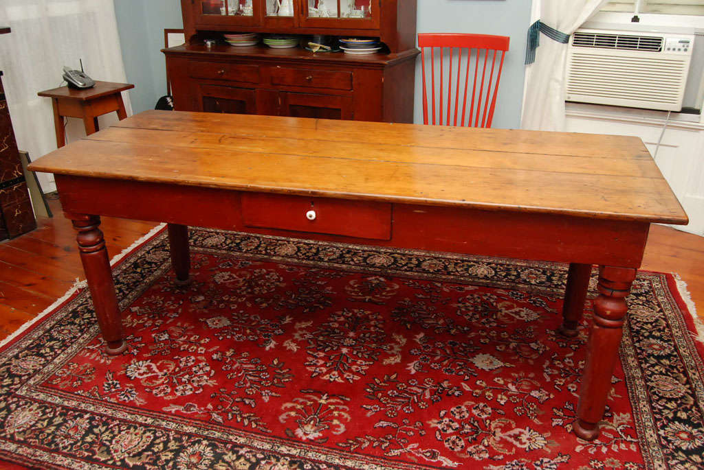 A lovely pine table once used in a as a store fixture with waxed three board pine top  on a red milk painted base with bold turned legs and a drawer in the apron.  Note the brass tack heads on the side of the table used to measure yard good.<br