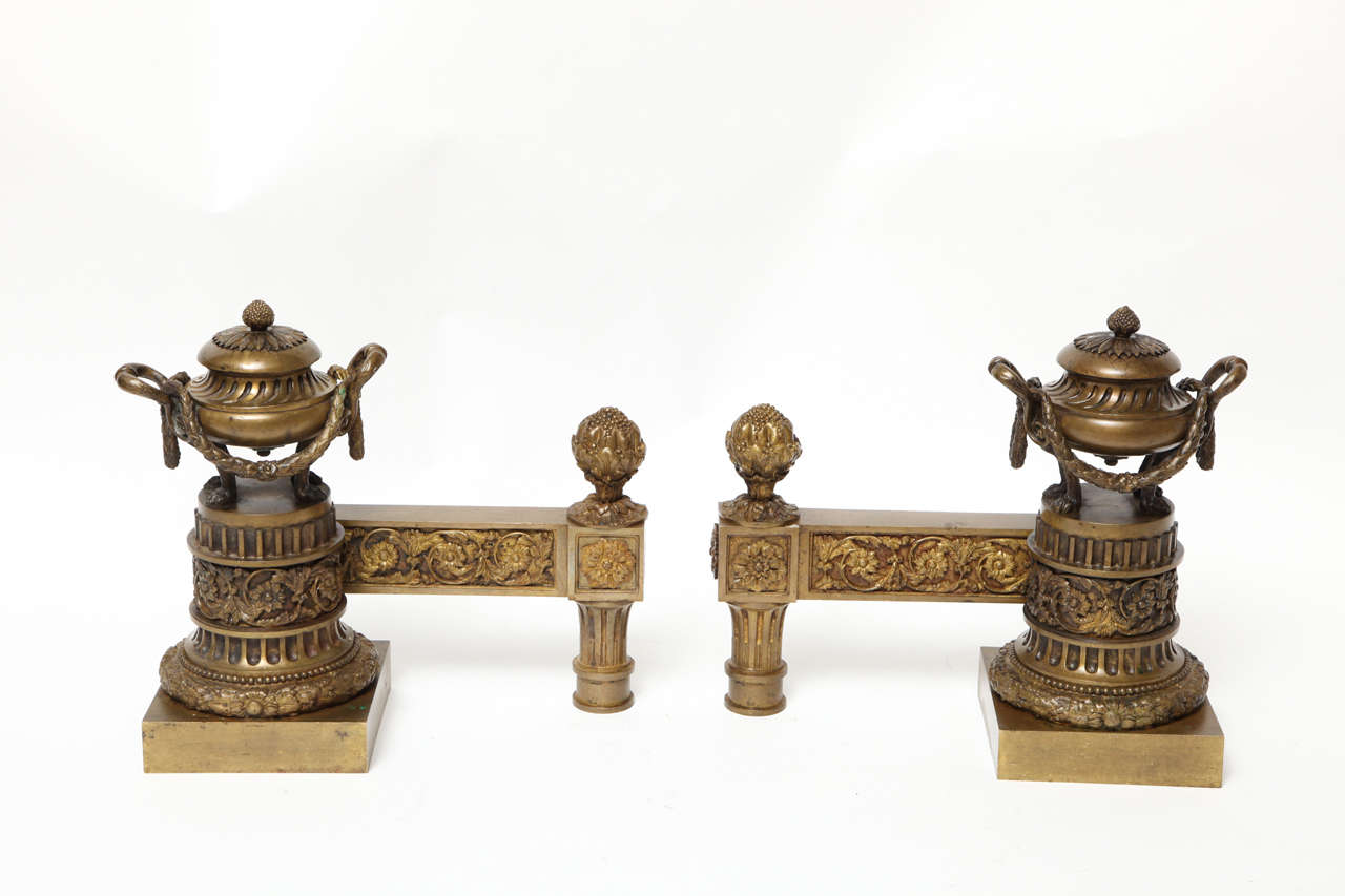 A pair of bronze French Louis XVI-style andirons, with amphoras supported by reeded columns with laurel wreath bases. Right and left-facing and with horizontal t-shaped supports designed to hold a firebasket.