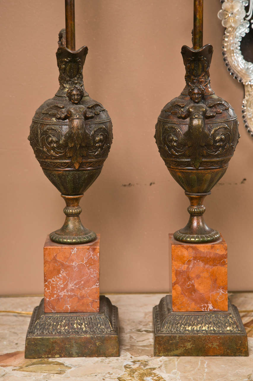 A pair of finely cast bronze ewers mounted as lamps. This neoclassical pair of turn of the century ewers both have curved handle holders leading to a full bodied woman mounted on a bolubus shaped ewer. Both are mounted on marble bases.