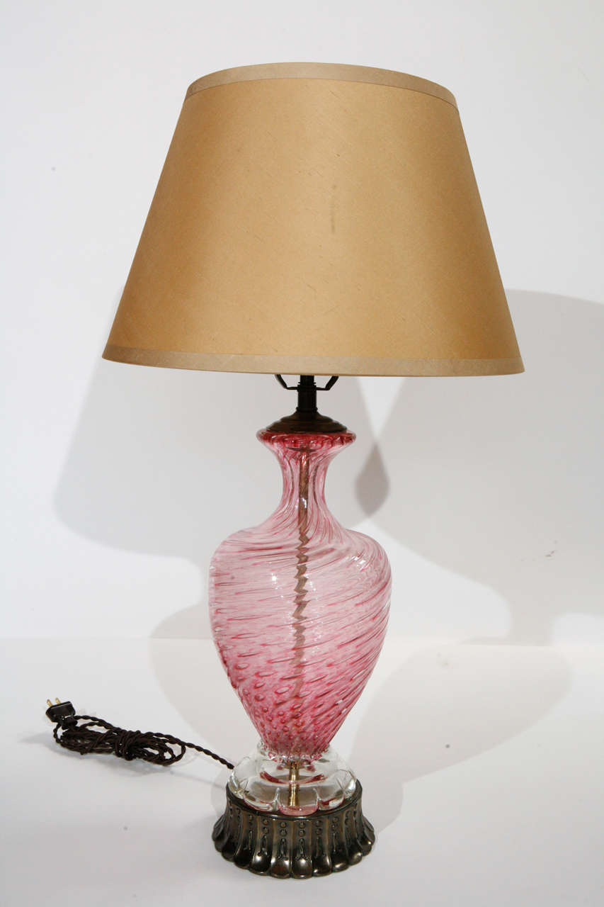 A vintage pink Murano lamp, decorated with swirl design and controlled bubbles. Base is an antiqued brass metal with an abstract design. Has a dark beige silk shade. Base is 6 1/2