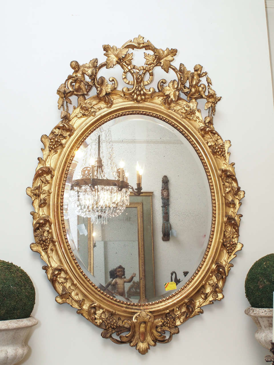 A beautiful oval carved giltwood mirror with trumpet flowers and grapes all around with two angels sitting on each side of the top of the mirror.