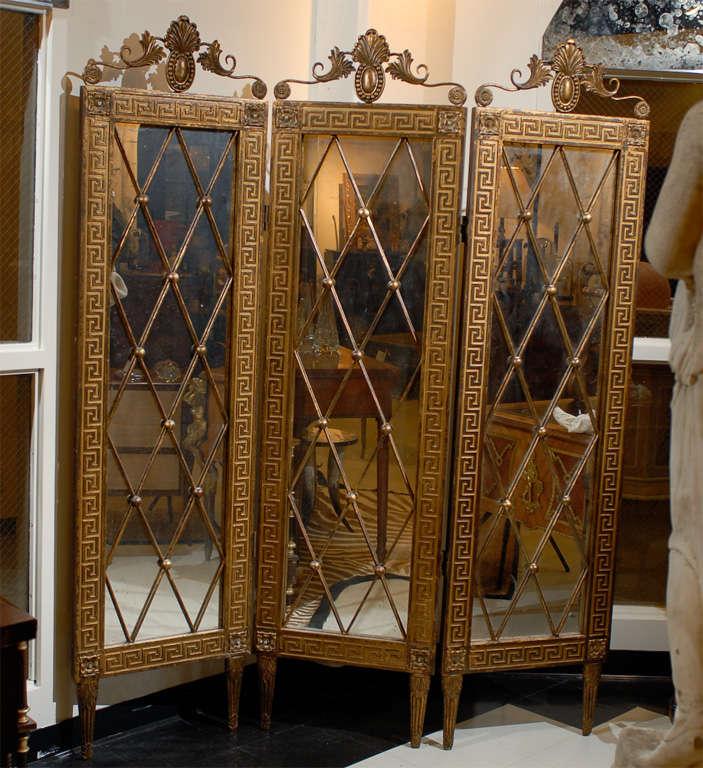 Hollywood regency gilded wood 3 panel mirrored screen with antiqued glass. Mirror divided by gilded metal diamond shapes with metal spheres at intersections. Classical carved wood Greek key runs the length of the top, bottom, and sides of panels