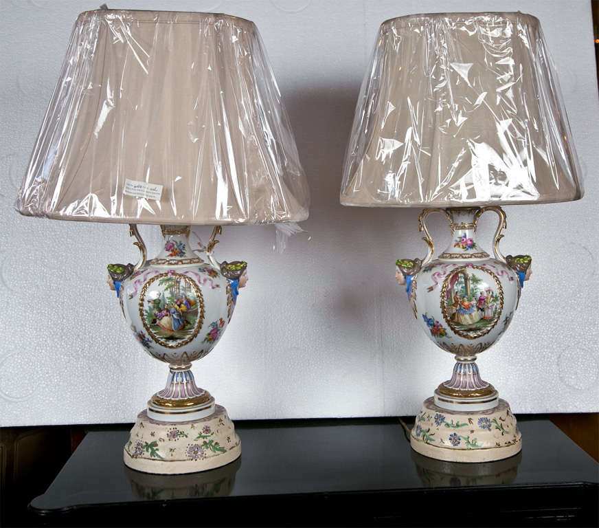 Pair of paint decorated porcelain lamps. Possible Meissen. The fronts depicting a finely done genre scene, the reverse with floral decorated scenes. Both having woman heads and handled sides. Previously urns now mounted as lamps each supported by a