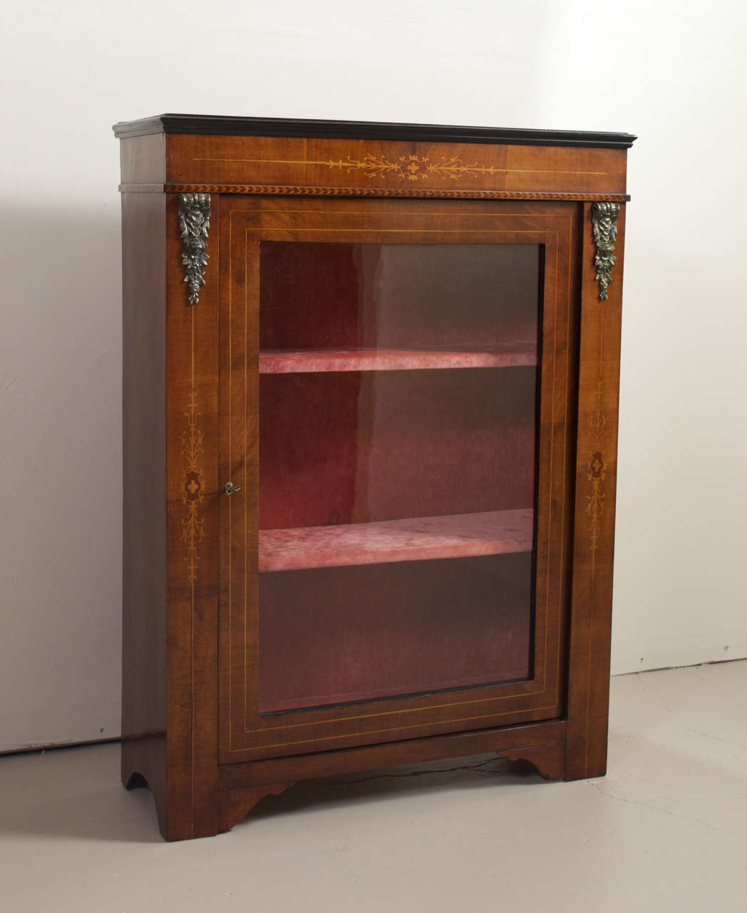 A beautiful inlaid French walnut side cabinet with brass ormolu. This single glass door cabinet has a red velvet lined two shelf interior. There is a black ebonized molding around the top and an elegant satinwood inlay pattern below that also goes