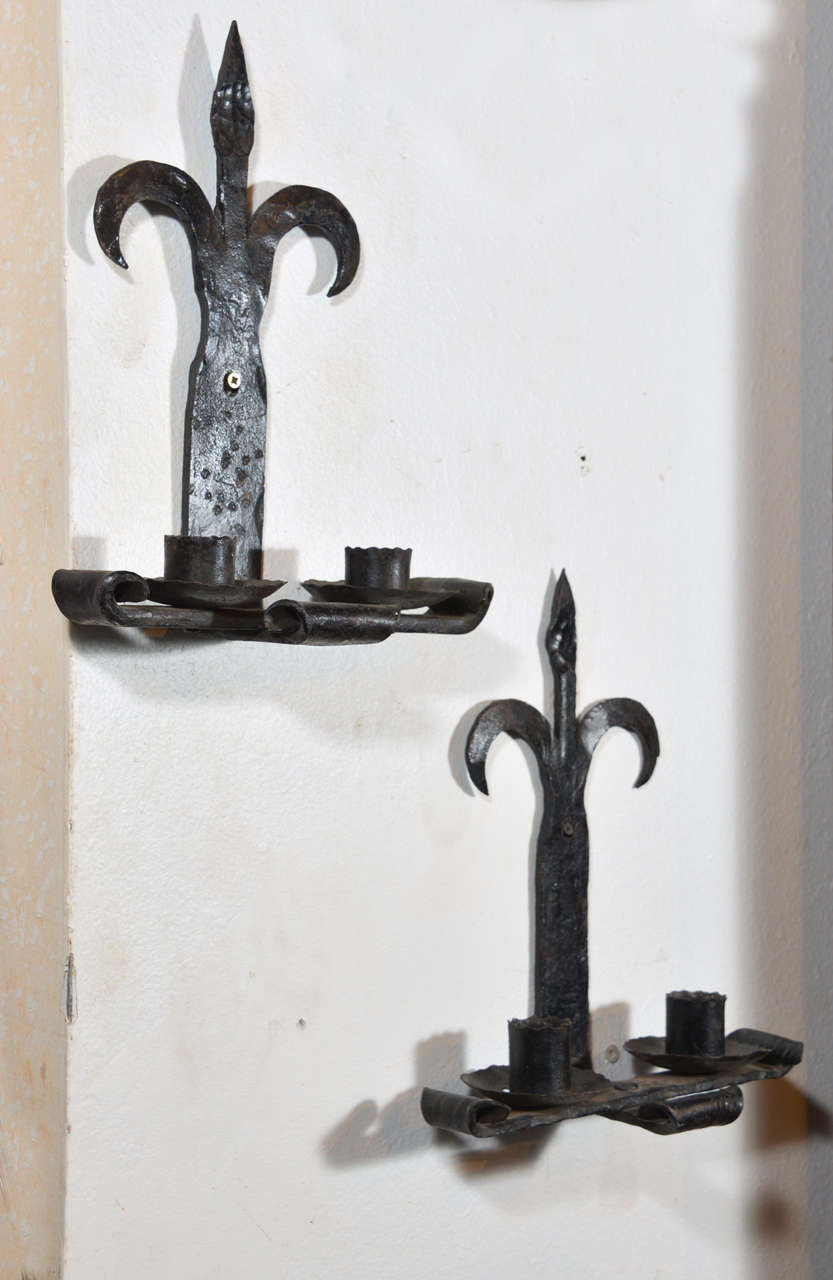 This pair of antique hand forged iron fleur de lys sconces from France has scrolling motifs throughout.  The back plate has a fleur de lis motif at the top and extends down onto a platform of three scrolled bands.  Finally, there are two candle cups