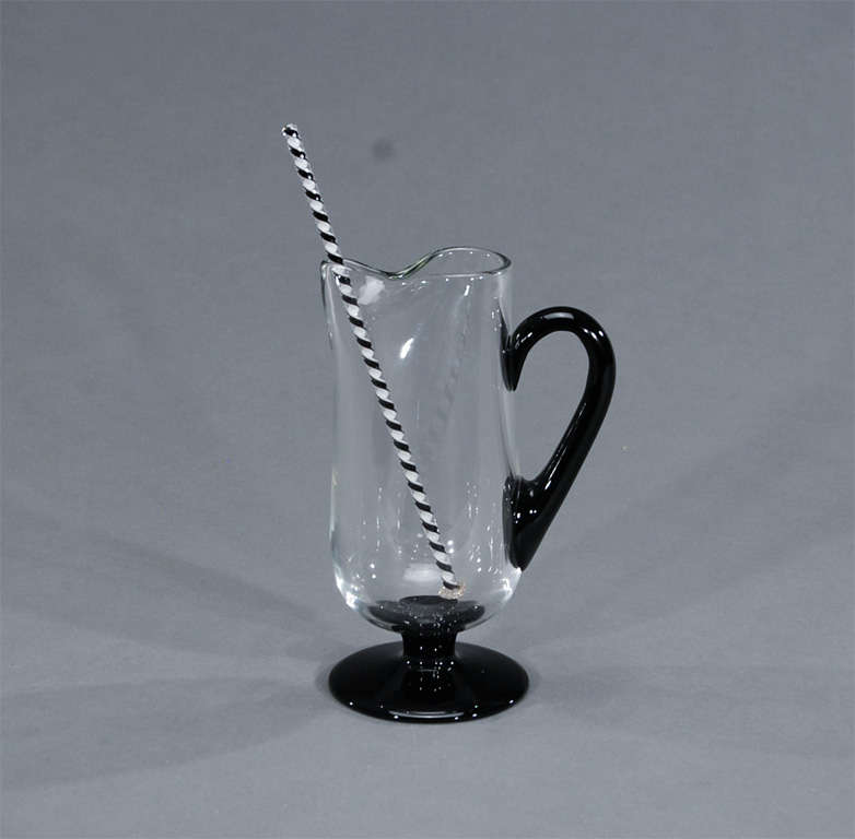This handblown crystal martini pitcher with applied black crystal handle and base provides a useful piece with an artistic and decorative statement. The accompanying stirrer is a two-color rod with ribbon-like effect, cased in clear called the