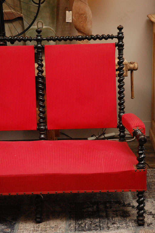 A settee or sofa with an ebonized barley twist frame with a raised bobbin and finial back crest, barley twist front  legs, and padded armrests with finial ends. In a red raised stripe upholstery with studs