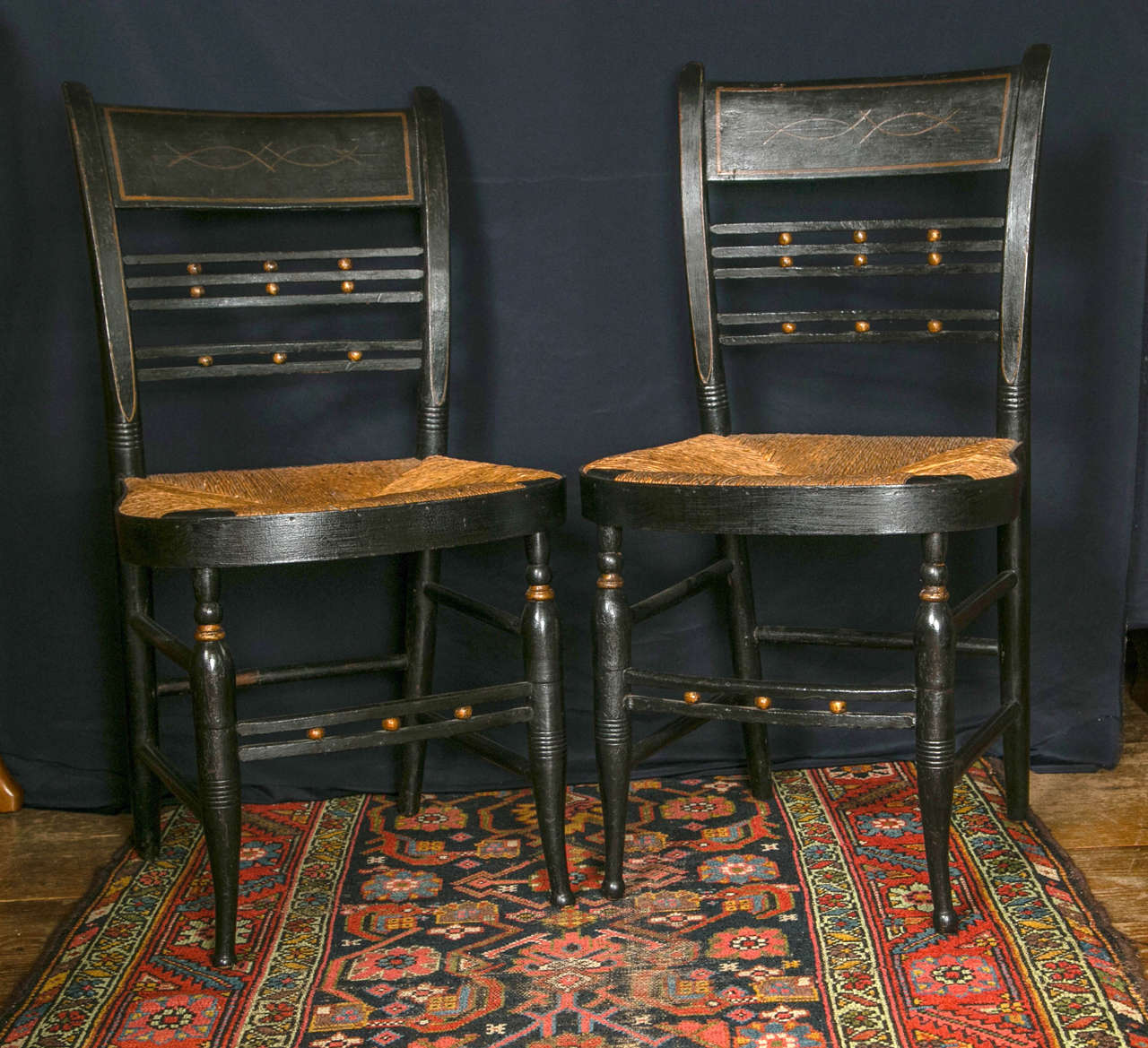 Black painted with gold accents, these rush-seated chairs aspire to grander environs, yet their humble stature will always foil a much wished for trip to the parlor. A late 19th century version of hall chair, they were meant to weather the drafts of