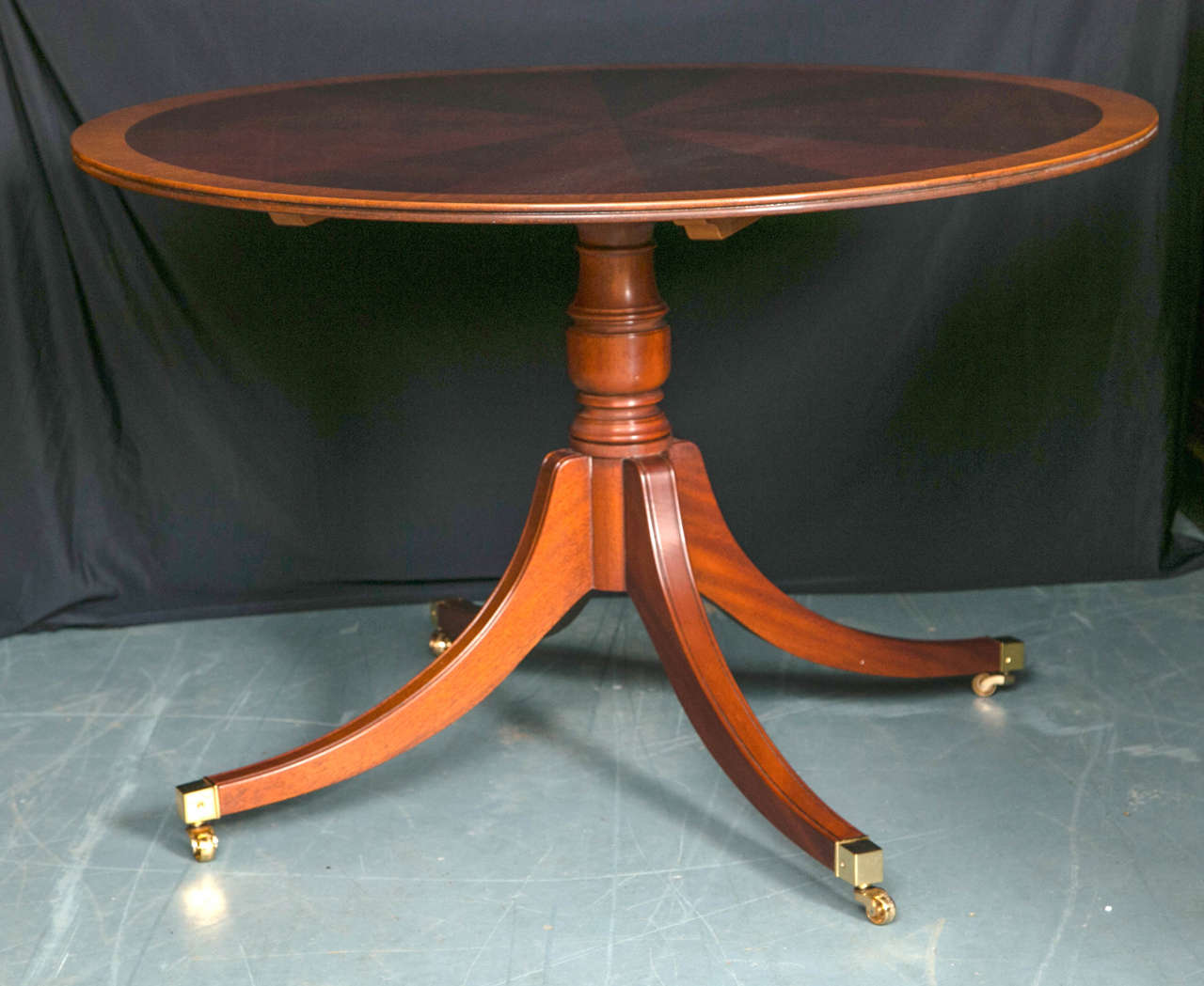 Created in the style of tables from the 18th century, this circular, mahogany starburst table is supported by a single column pedestal with four splay legs ending in brass toe caps and casters. Banded in satinwood and having a reeded edge, this