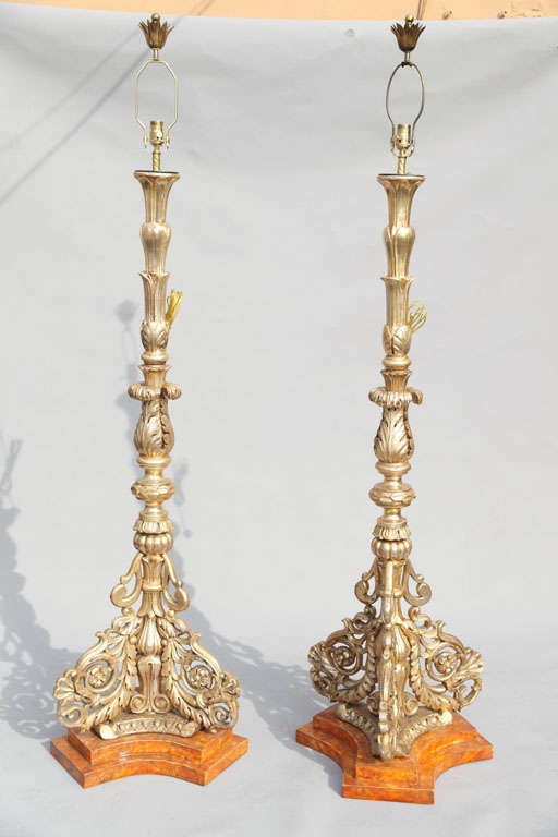 Lavish pair of prickets, now fitted as floor lamps, of carved silver giltwood with acanthus and laurel leaf designs, each raised on tripartite base with scrolls and rosettes, on later conforming custom faux painted plinths.
