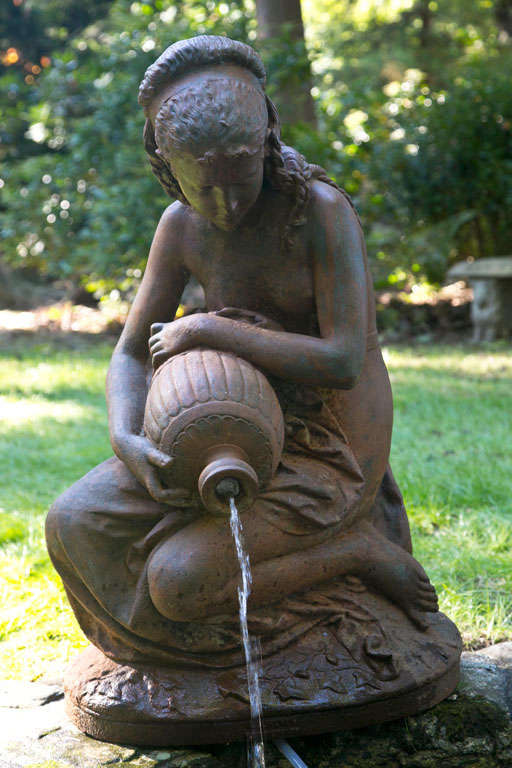 A cast-iron fountain figure of a seated girl with serene expression, her hair meticulously rendered with braids, the overturned jar on her knee plumbed for water, marked 