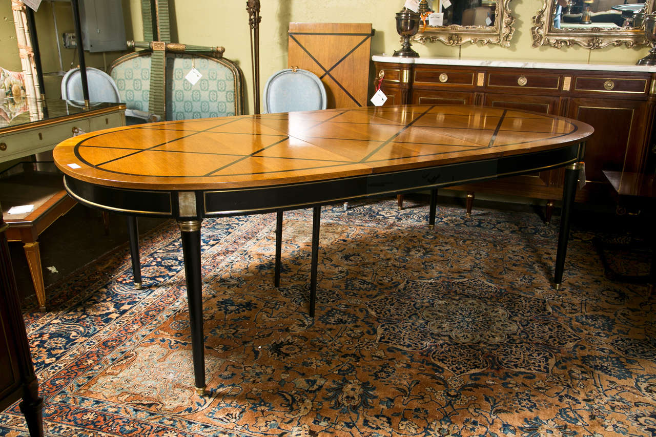 French Louis XVI style two-leaf dining table, parcel-ebonized, the oval top with a parquetry pattern, atop a banded narrow frieze decorated with bronze mounts, raised on circular tapering legs ending in capped feet. By Jansen.

Two leaves