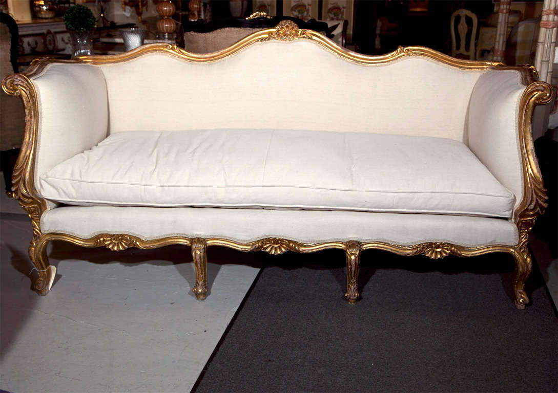 Beautiful French Rococo style giltwood canape sofa, circa 1920s, elegant carved frame in original gilding, scalloped back centered by a shell crest, rolled arms, upholstered in linen and burlap exterior, cushioned seat, raised on cabriole