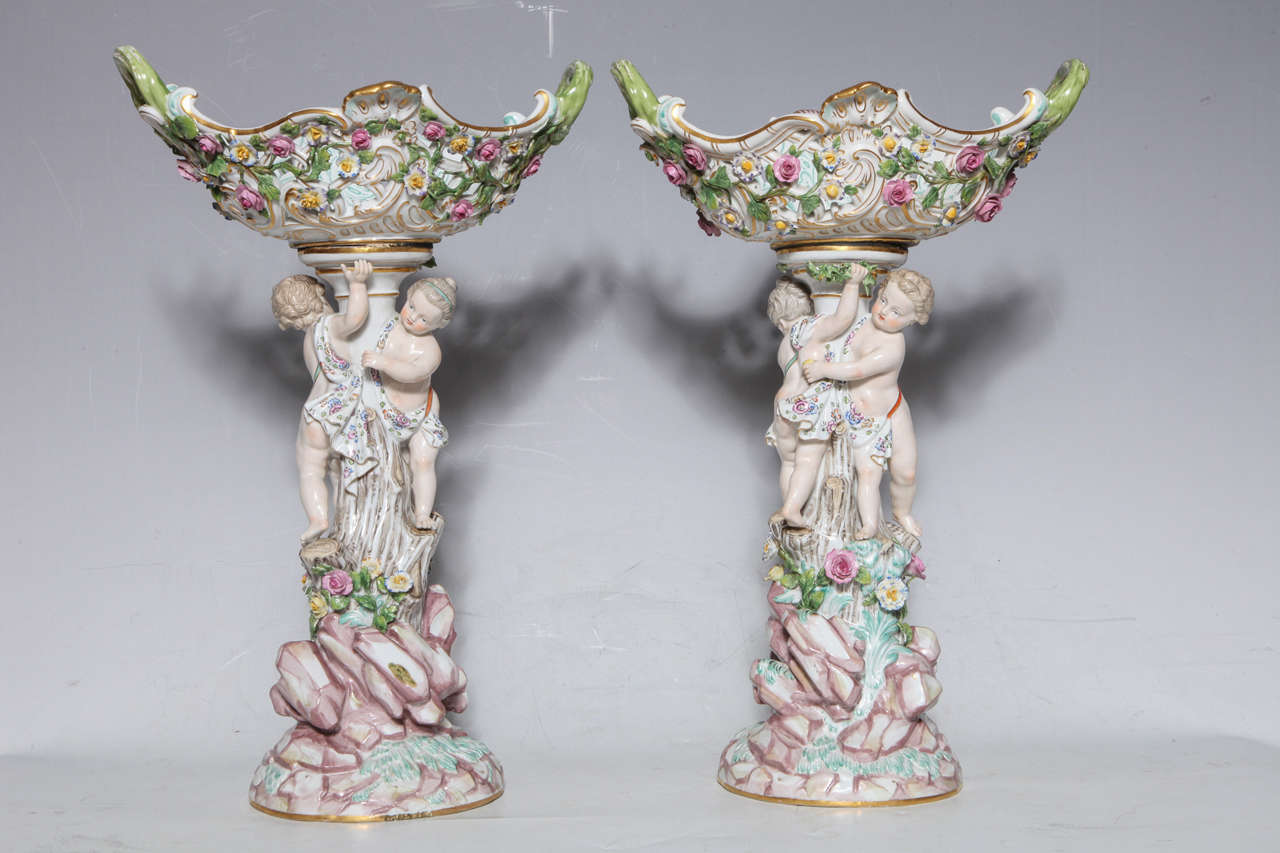 Pair of 19th century Meissen Porcelain compote centerpieces with cupids running after each other around a tree with filigree baskets on top with raised flowers and vines. Hand-painted with beautiful colors, first quality Meissen, blue under glazed