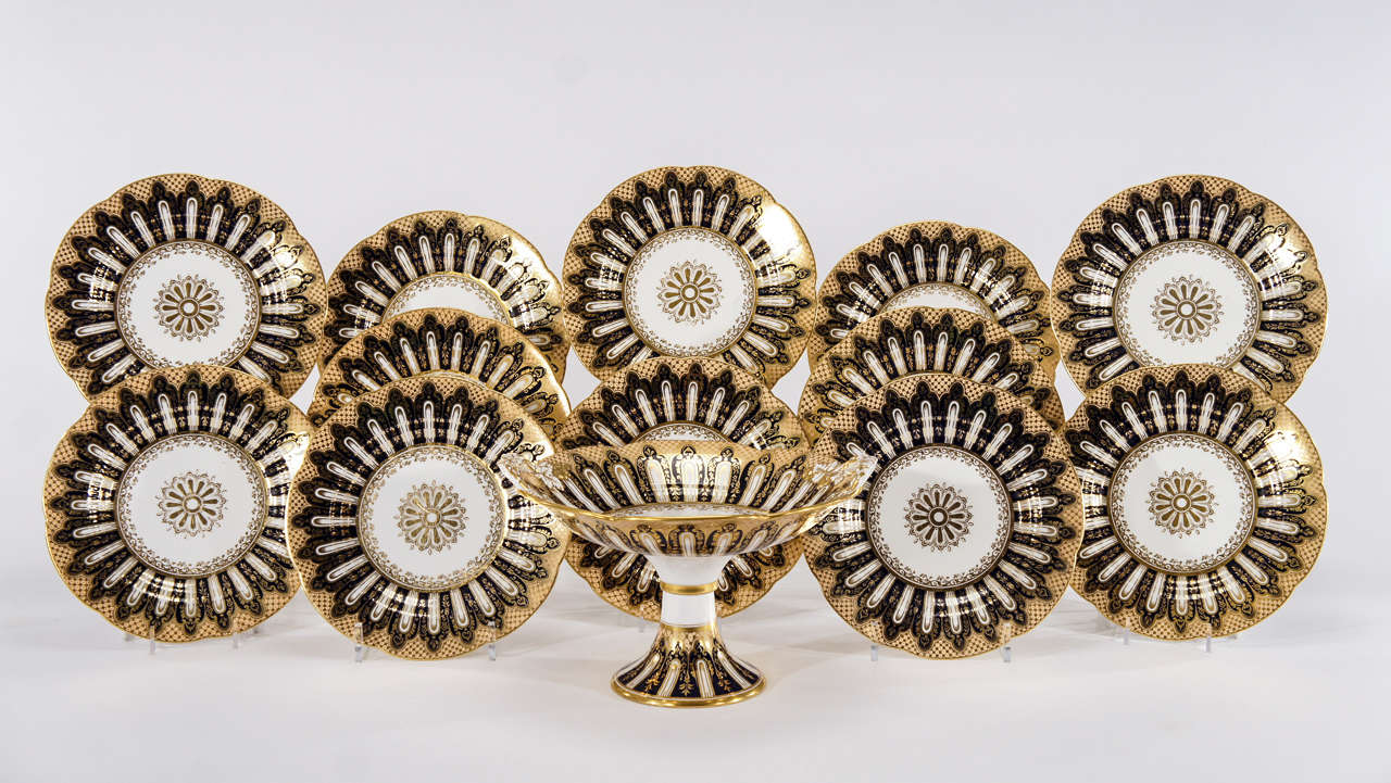 This is a lovely 19th century set of 12 extravagantly decorated dessert plates and matching footed compote with molded leaf handles trimmed in gold.
With a bold graphic design, these incorporate a deep blue, caramel, white and gold palette with a