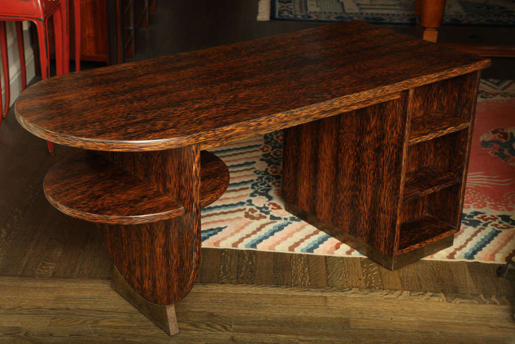 This palmwood and oxidized brass desk has four pedestal drawers on one side and two adjustable shelves on the other. The top is rounded on one end with a round shelf below on the exterior and interior side. It can be used protruding from a wall as