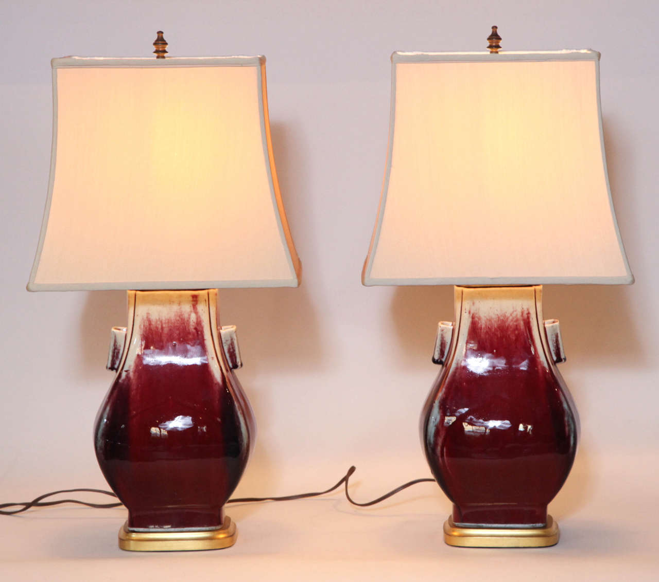 Rectangular with tapering neck and two tubular handles. Oxblood red glaze streaks with corners in purple. Mounted on a wood base and wood top. Both entirely covered with gold leaf. Rectangular silk shades.

Electrified for 3 way bulb and in good