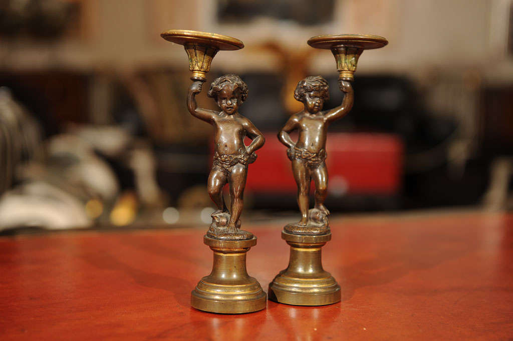 Each putti-form depicted with delicate features, an arm outstretched to support a candle cup and standing on a pedestal base.