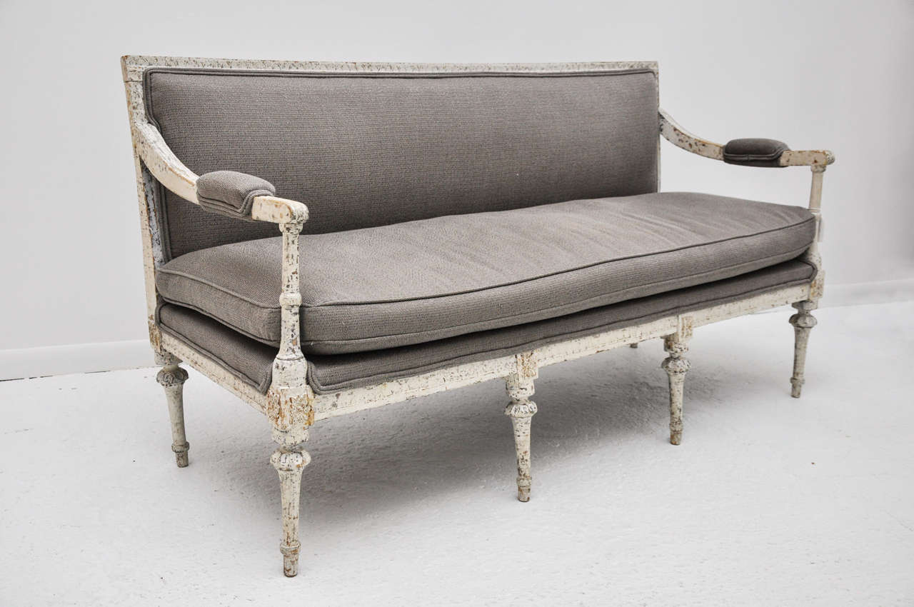 Carved & painted wooden settee in Gustavian style. Smooth, unfluted inverted-trumpet form legs with reverse-gadrooned tops, leaf-carved top rail, applied composition paterae above legs. Finish is old white paint, scraped back to reveal undercoat &