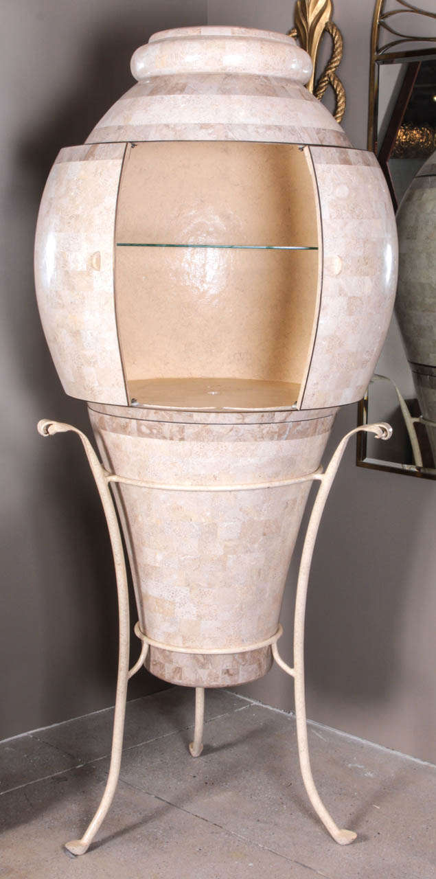 A rotating circular bar cabinet with integral sliding doors is set on a painted iron tripod base. The interior is illuminated by an oculus on the top. The body is entirely covered in warm beige fossil stone tesserae, with bronze trim. By