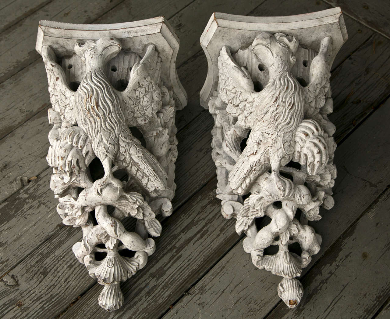 A true pair of brackets with one  eagle(?) facing left, the other facing to the right.  Out spread  wings help hold up the concave sided shelf.  The birds set among branches and foliates. A single coat of worn white paint covers the brackets. The