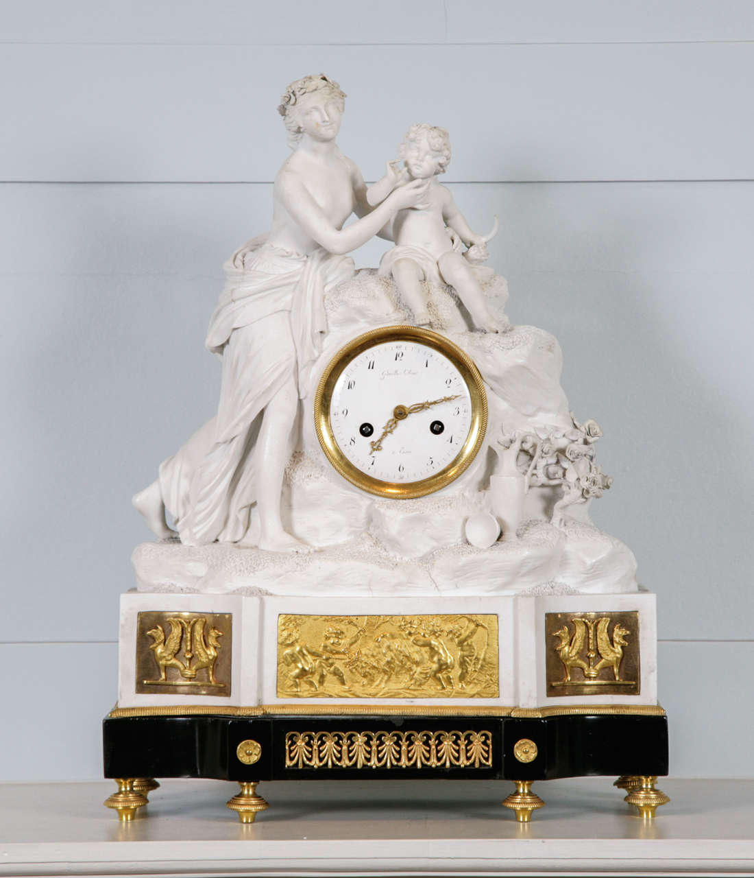 A substantial late 18th century French mantel clock by Gavelle of Paris.  The sculptured case of bisque porcelain depicting a classical female standing figure, with a winged cupid, sitting on rocky boulders of bisque porcelain, with a vase and
