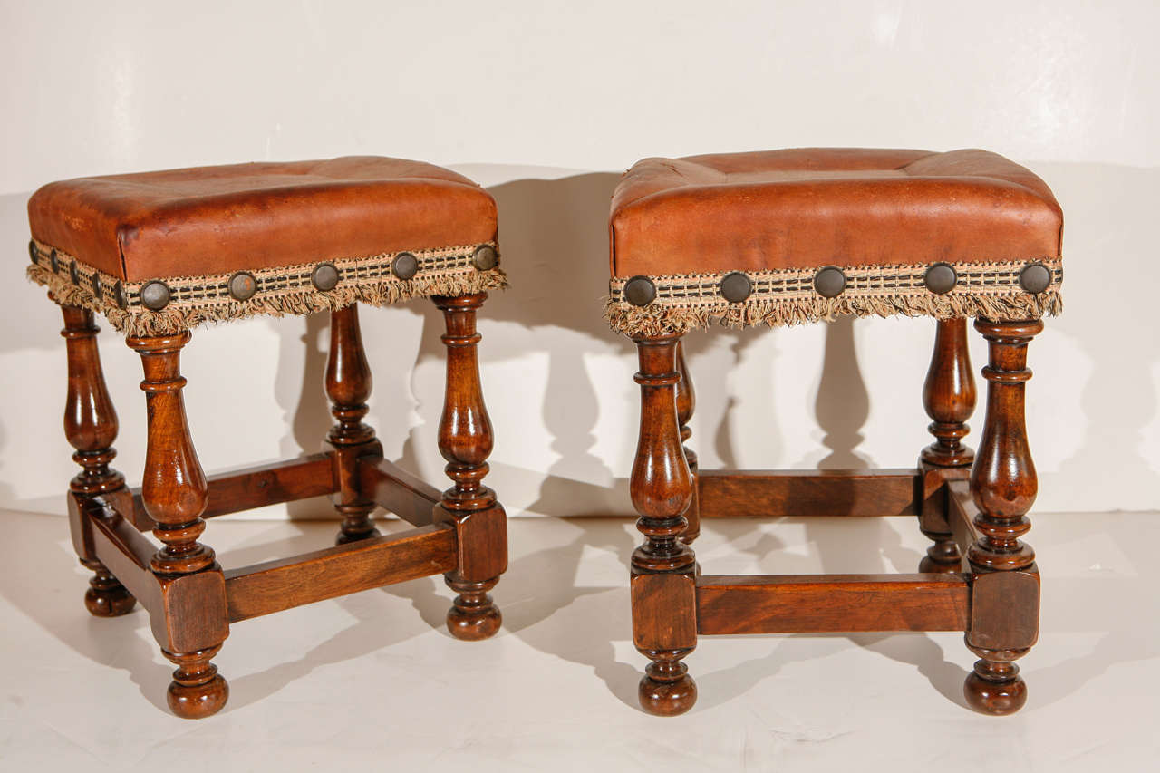 Pair of turned, Italian walnut footstools with leather upholstery, oversize nailheads and fringe trim.