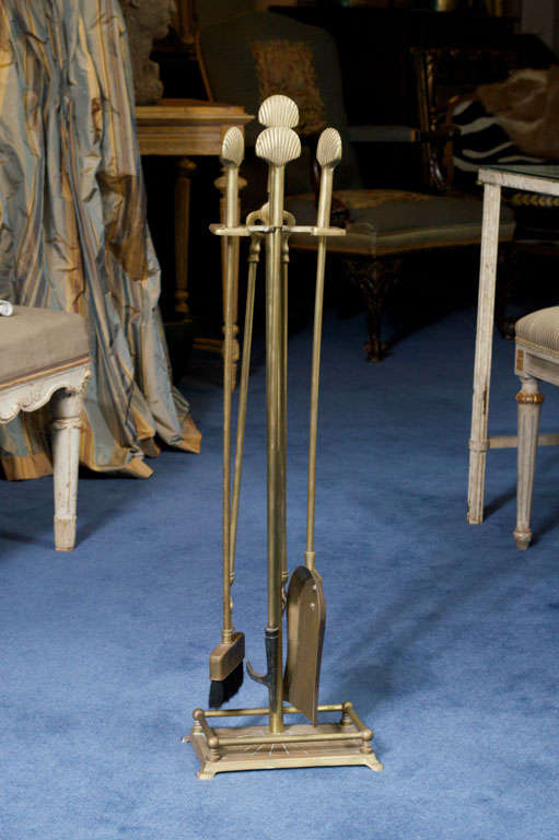 The assembled group of  elegant and sleek brass tools comprising of a small broom with shovel, poker and pair of tongs, each with shell-molded handles, including stand with hammered base and brass rails ending on shell feet.