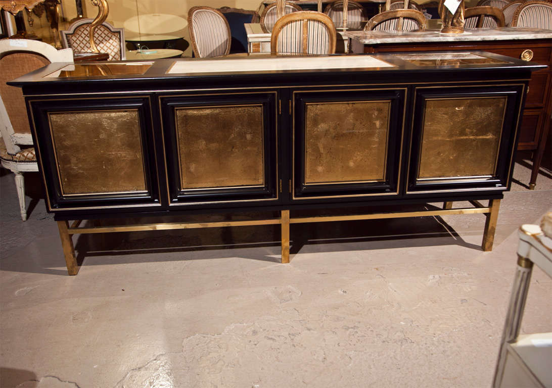 A mid-century style ebonized sideboard, 1960s, beige marble in the center flanked by gilt-glass tops, over a conforming cabinet with gilt-glass doors, open to shelving interior, raised on bronze base. By Maison Jansen.