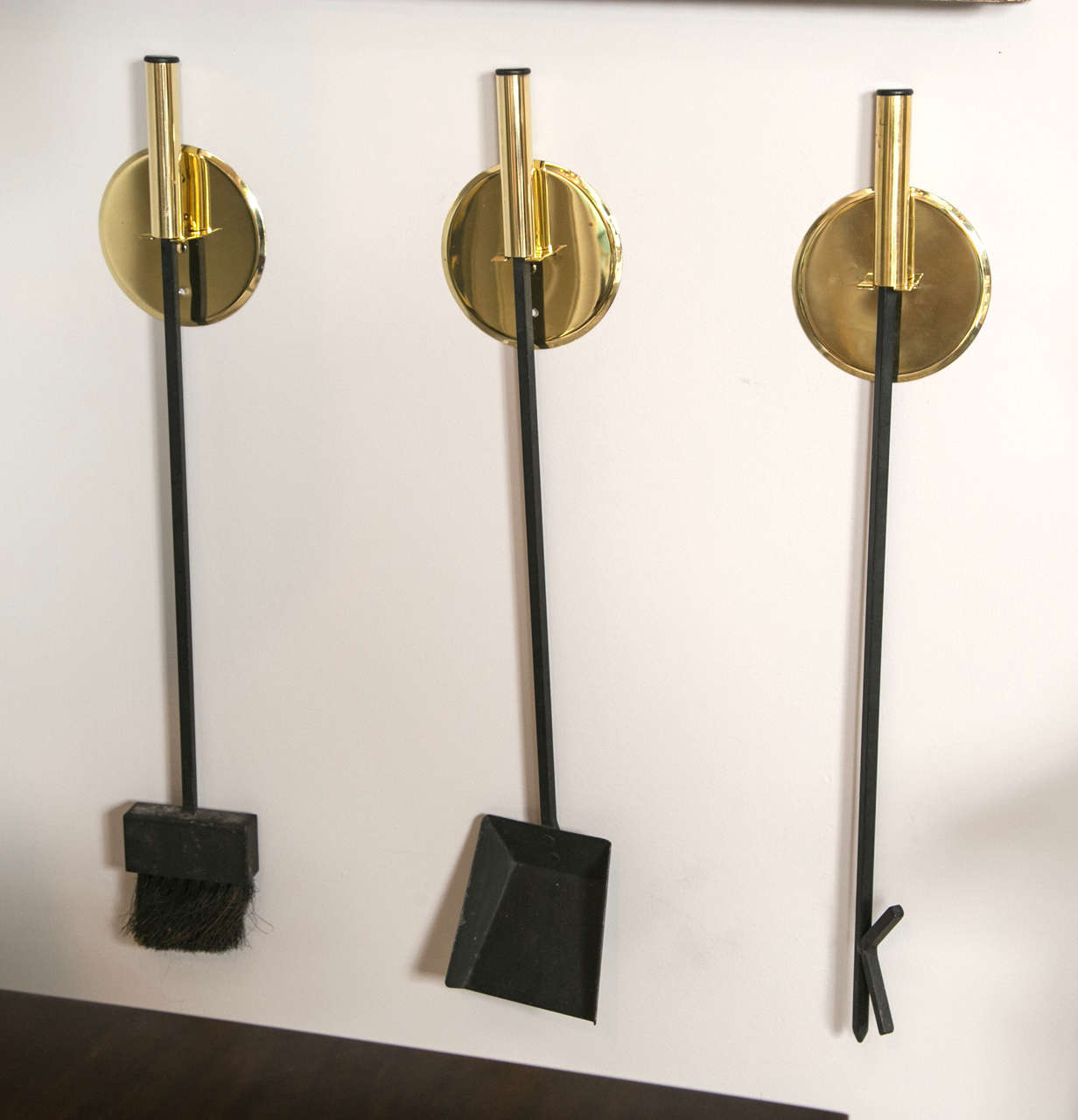 rare set of three brass Mid-Century fireplace tools, comprised of  3 discs brackets that mount to wall, each separately holding a fire tool. Each blackened iron fire tool (poker, brush and shovel) has a brass handle. Newly polished.