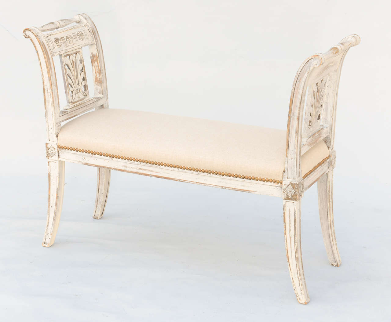 Narrow bench, in Neoclassical taste, having a painted finish showing natural wear; its rectangular stuff-over seat upholstered in linen with nailheads, flanked by out-scrolled arms with pierced anthemion splat, finished by turned handles, raised on