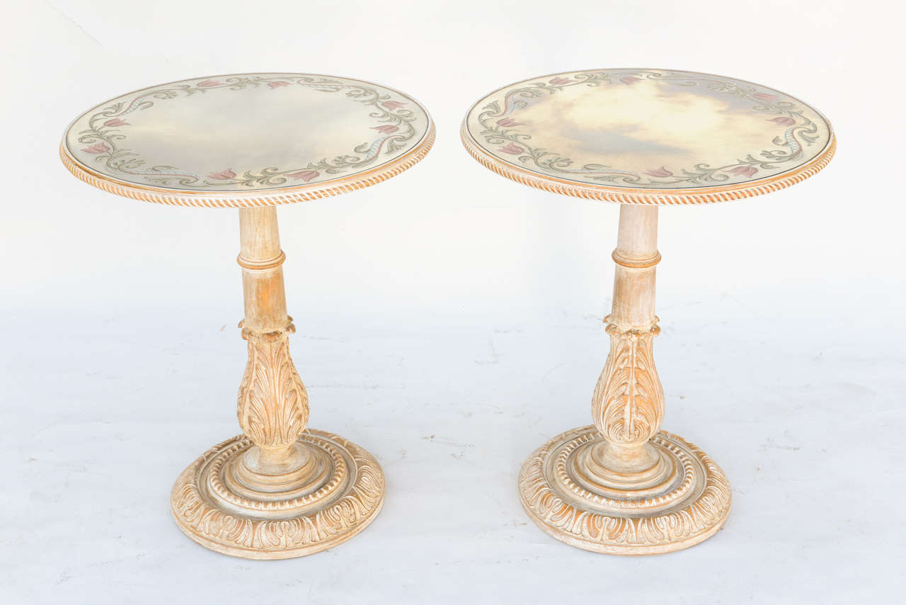 Pair of tables, each having a round mirrored top with reverse painted floral vine decoration, surrounded by gadrooned border, raised on pedestal stands carved with acanthus, on round foot base.