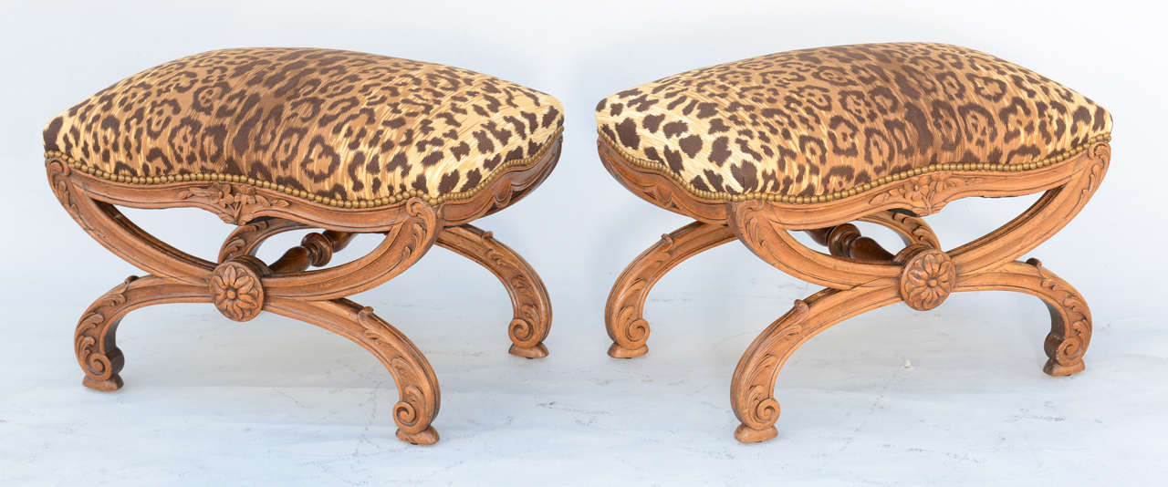 Pair of benches, or ottomans, each having a stuff-over seat upholstered with nailheads, on channeled X-frame base, its serpentine seatrail carved with flowers, raised on X-form legs carved with acanthus and scrollwork, joined by turned stretcher,