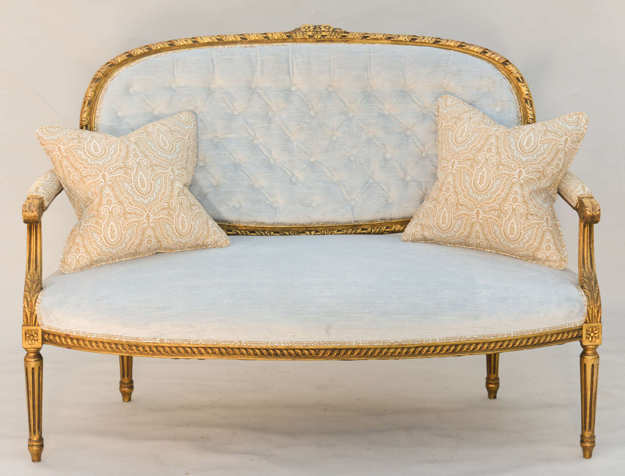 Louis XVI giltwood settee, having a tufted oval back in gadrooned frame surmounted by carved flowers, padded armrests on channeled uprights, stuffover seat on bowed rail with complimentary carving, raised on round fluted tapering legs, ending in