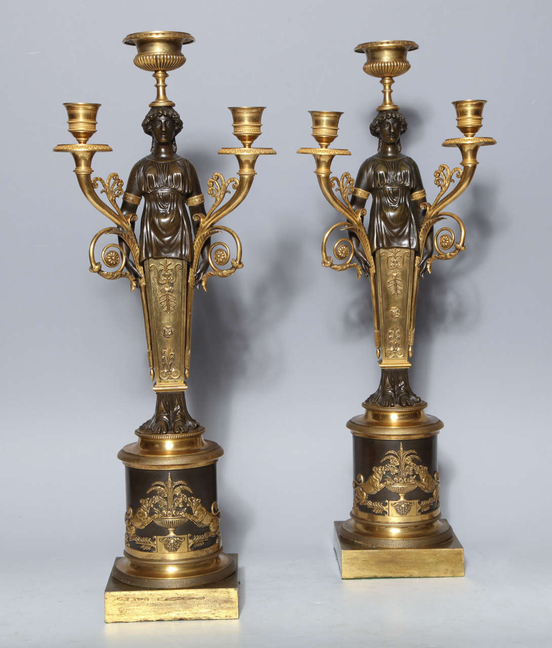 A fine pair of antique French Empire period two tone, patinated and dore bronze figural candelabra. Each modeled as a neoclassical standing maiden holding a branch in each hand with wreaths and palmets. Each base adorned with a pair of lions