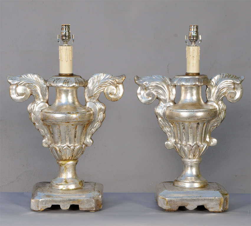 Pair of lamps, of carved silver giltwood, each a porta palma base; a fluted urn with large scrolling handles, on leaf-carved socle, raised on round foot and square plinth. Lamped, with French-style wiring.
