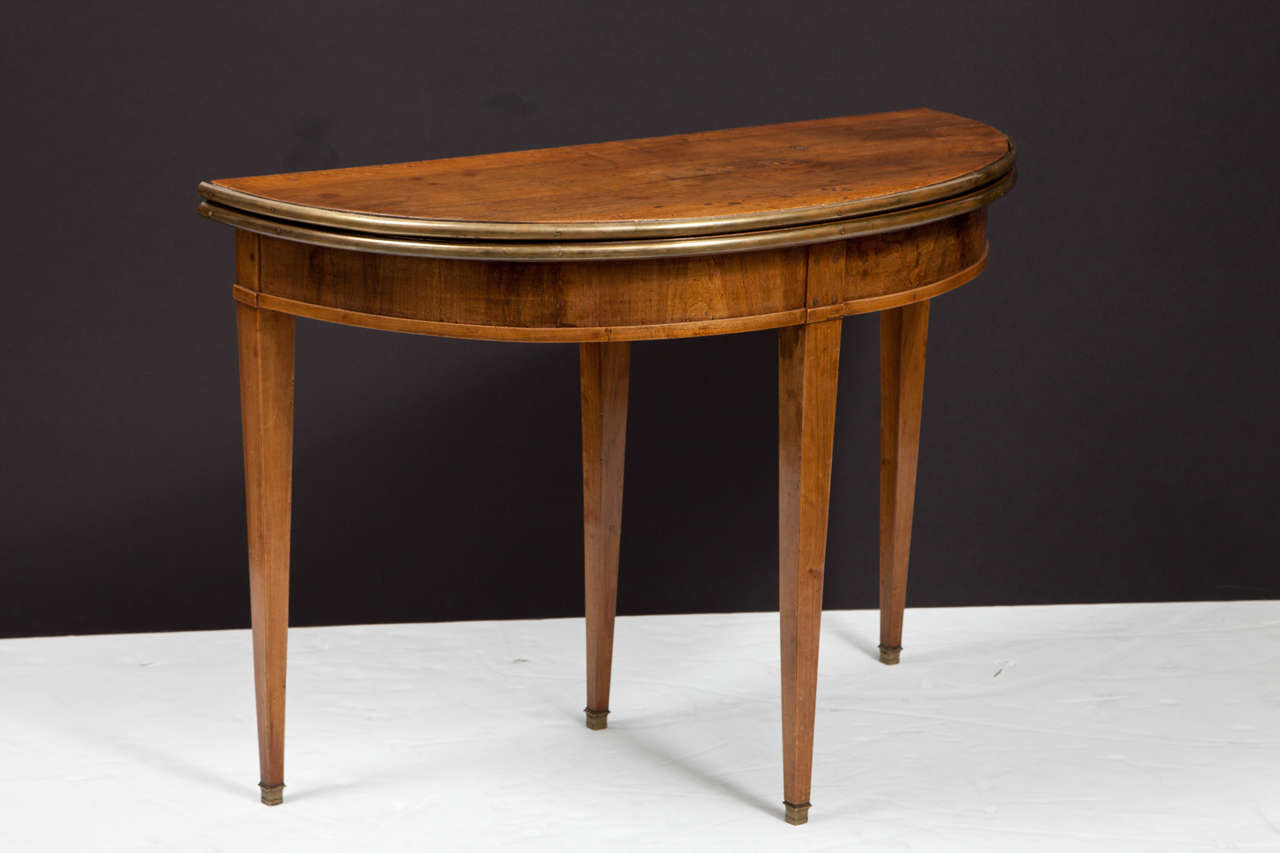 The brass banding and warm patina of this demilune table give it great character. In light mahogany, the table has a gate leg and opens to a full circle, raised on tapered legs with brass sabots. The table is 19